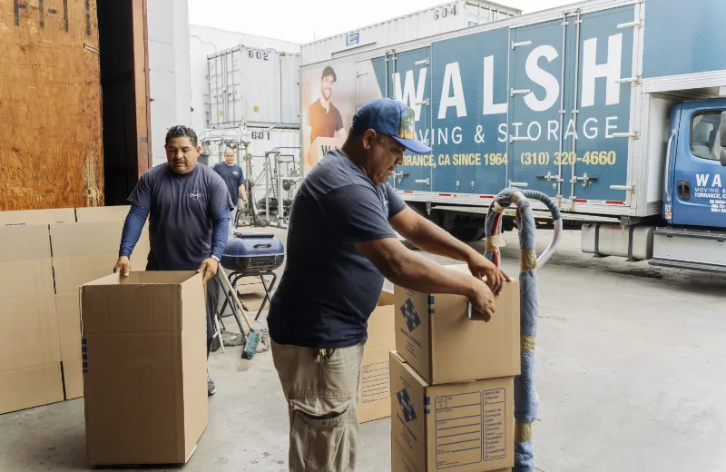 Two workers assemble cardboard boxes outside a Walsh Moving & Storage truck in a warehouse area.